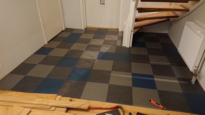 Nyinstallerat schackrutigt golv i en hall med trätrappa, verktyg syns i förgrunden.