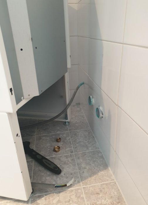 Öppet grått skåp med synliga vattenanslutningar och avloppsrör i ett delvis kaklat badrum, verktyg på golvet.