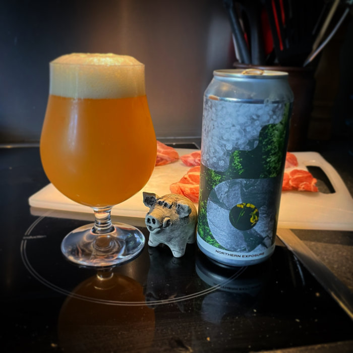 Glas med öl bredvid en ölburk, liten grisfigur framför, skärbräda med lax i bakgrunden.