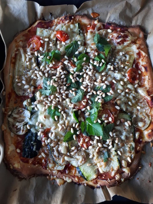 Hemlagad pizza toppad med tomater, zucchini, ost och pinjenötter på bakplåtspapper.