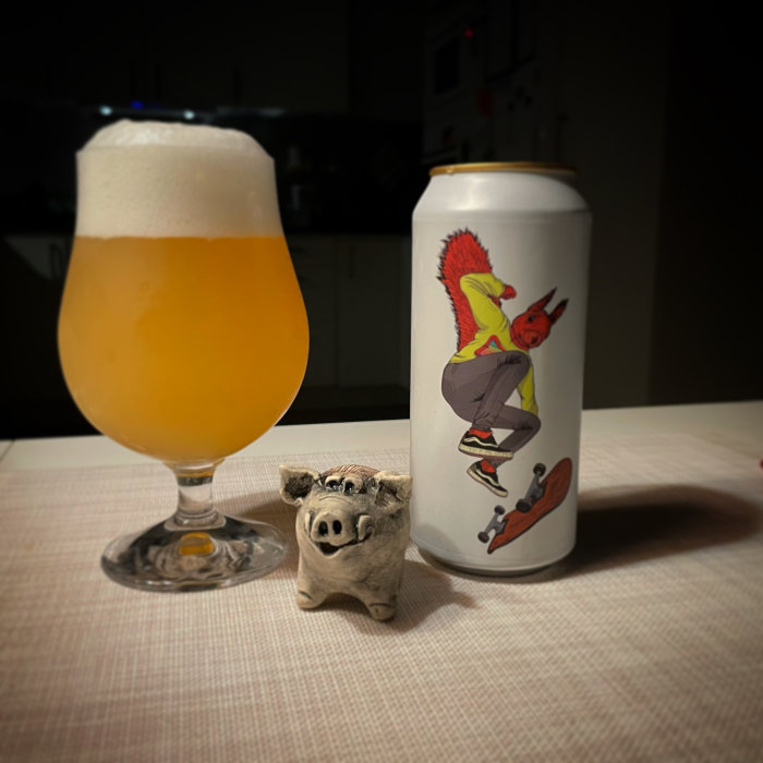 Ölglas med skummande öl bredvid burk med tecknad ekorre på skateboard och en liten grisskulptur framför.