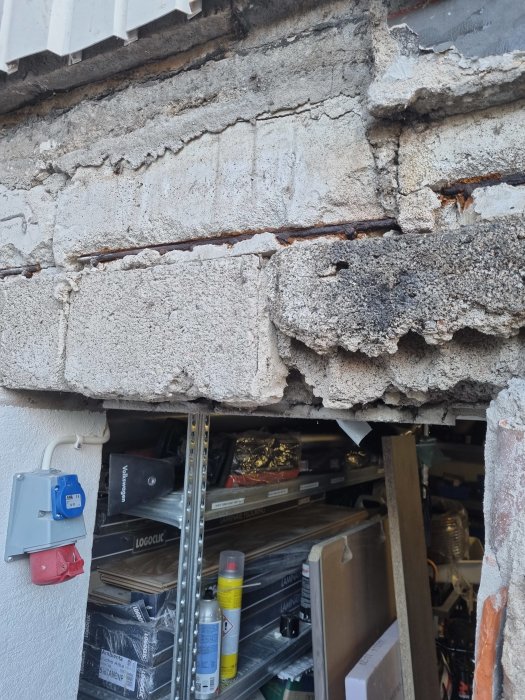 Bild av en skadad betongvägg med synlig armering och sprickor över en garageingång.