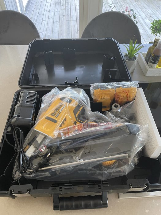 Ny Dewalt verktygssats inuti en öppen låda på ett bord, fortfarande i plastförpackning, med laddare och extra tillbehör.