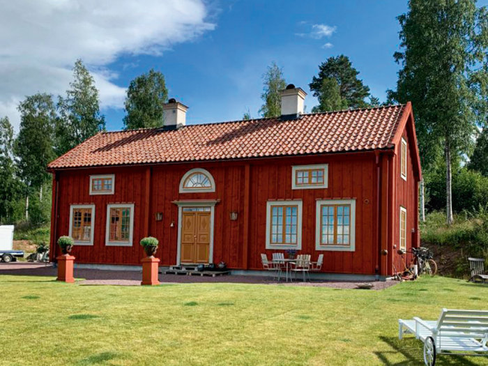 Rödmålat trähus med tegeltak och vitmålade fönster, omgivet av grön gräsmatta.