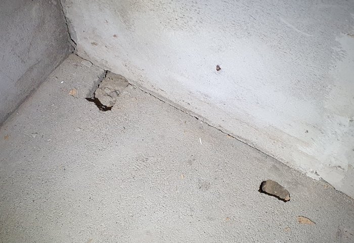 Två hål i ett betonggolv med synliga skador och vittring nära en vägg, vilket indikerar fuktproblem i en källare.
