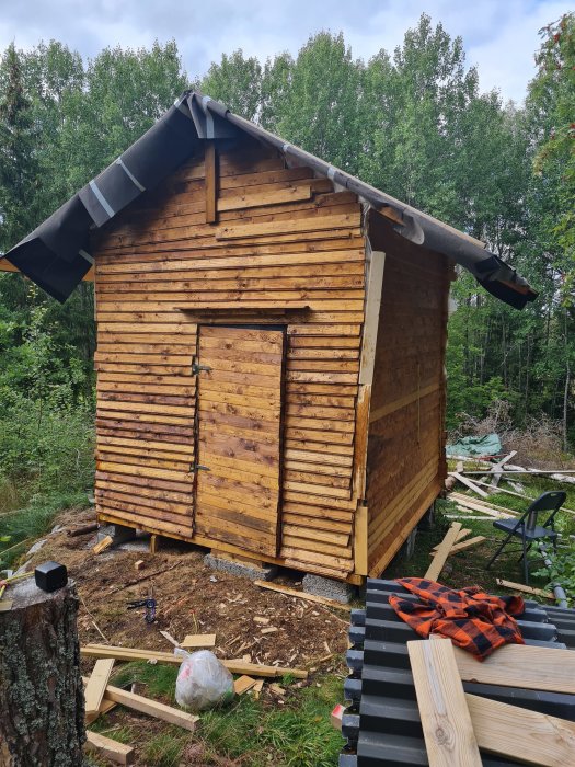 Nästan färdigställd träskjul med nytt tak och oputsade brädor i skogsmiljö, byggmaterial synligt i förgrunden.