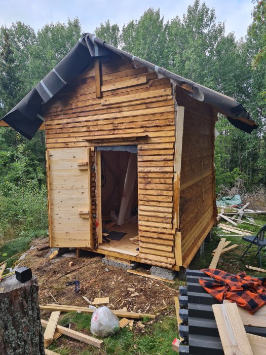 Nästan färdigbyggt litet träskjul med öppen dörr på en arbetstomt med skogsomgivning.