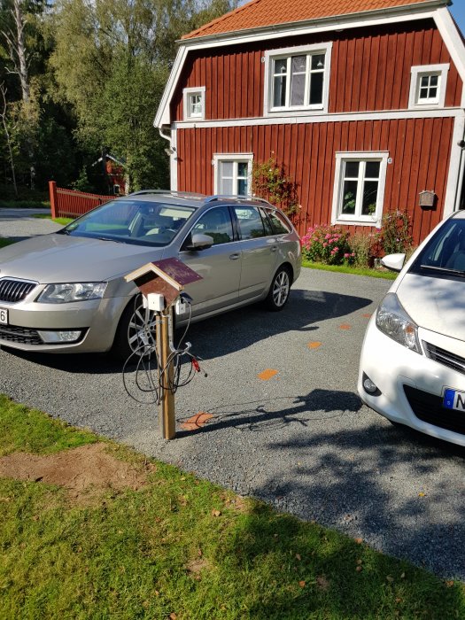 Enklare trästolpe med kopplade elektriska motorvärmarsladdar framför bilar, nära ett rödfärgat hus.