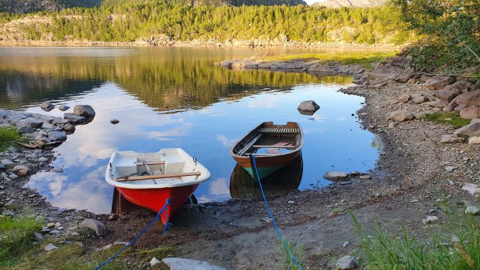 Två små båtar vid en sjöstrand, omgivna av skog och klippor, spegling i vatten.