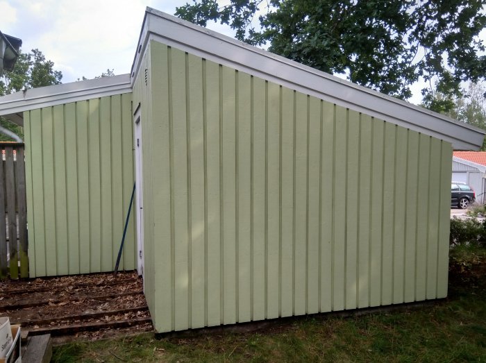 Grönt garage sammanbyggt med ett vitt förråd mot en bakgrund av träd och en klar himmel.