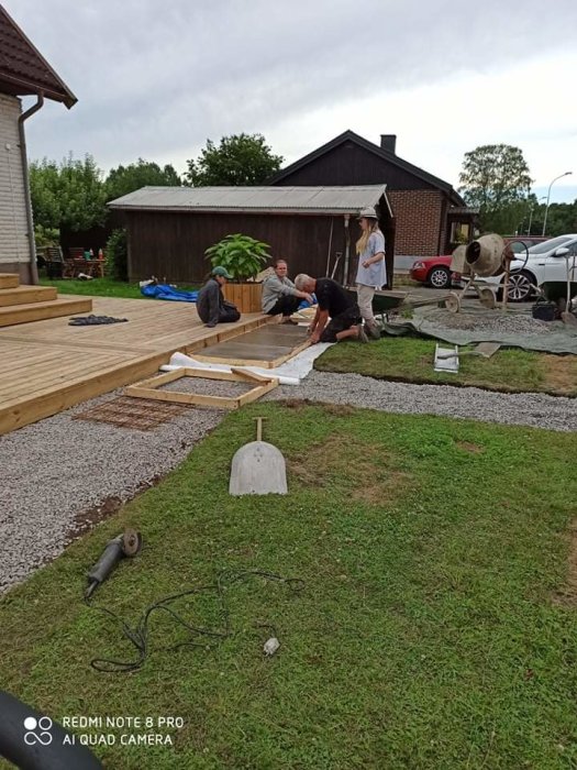 Personer som jobbar med att bygga en träaltan utanför ett hus, verktyg och byggmaterial synliga på gräset.