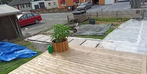 Färdigställt trätrall framför hus med krukväxt, områden med grus och presenning, samt röd bil i bakgrunden.