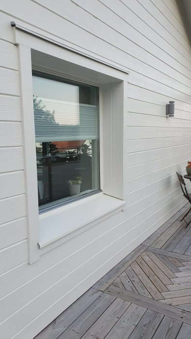 Ett fönster på en vit träfasad med persienn, intill en terrass med trägolv och en utomhuslampa.