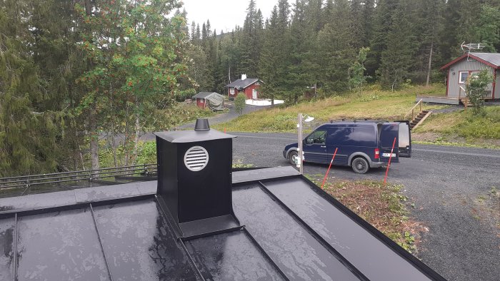 Svart takhuv monterad på ett glänsande plåttak med skog och röda stugor i bakgrunden.