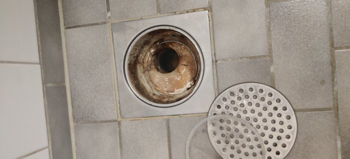 Golvbrunn med fastsittande filter och stålhandtag på kaklat badrumsgolv, nära vy.