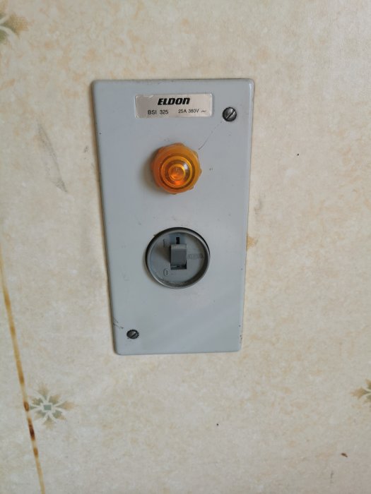 Gammal eluttag med etikett 'ELDON', orange lampa och strömbrytare på mönstrad vägg.