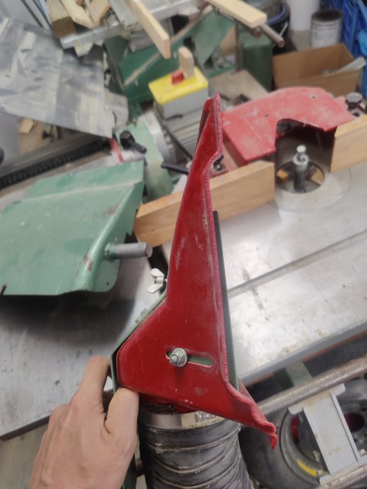 Hand som håller en röd verktygsdel i en verkstad med träbearbetningsmaskiner i bakgrunden