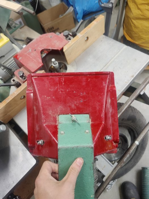 Hand håller grön verktygshandtag med röd metallkomponent i verkstad.