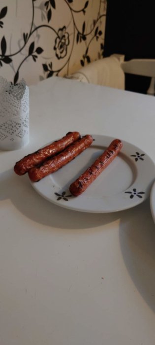 Tre grillade korvar på en vit tallrik med blomdetaljer på ett köksbord.