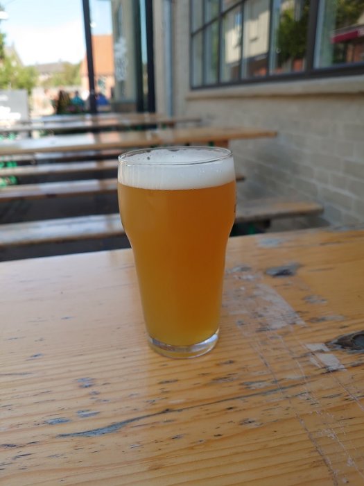 Ett glas öl av typen NEIPA på ett träbord utomhus, med suddig bakgrund av en gata och sittplatser.