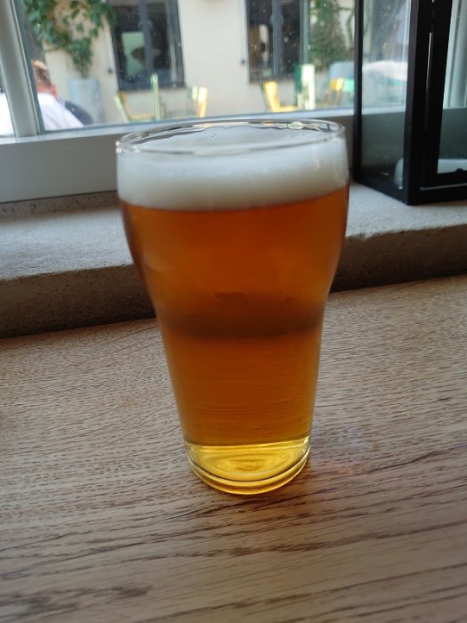 Ett glas öl med gyllengul färg och vit skumkrona på en fönsterbräda, bakgrunden visar en suddig stadsmiljö.