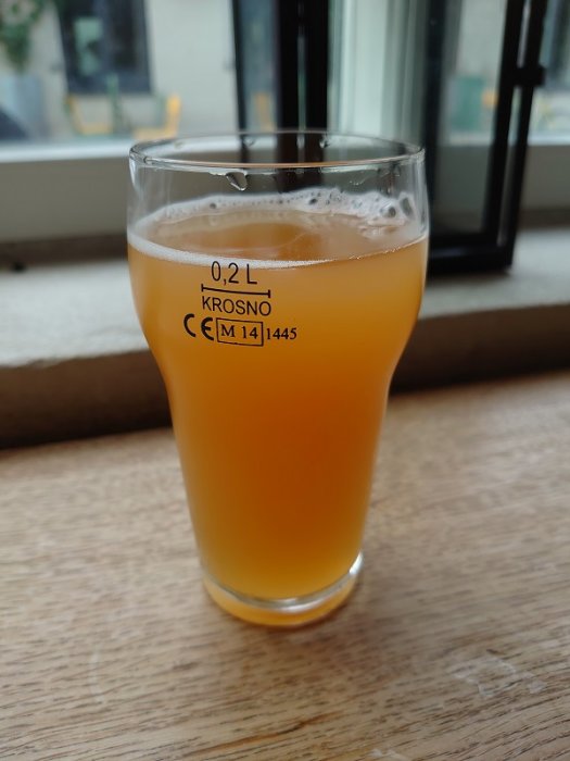 Glas med grumlig öl framför ett fönster, märkt 0,2L Krosno.