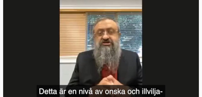 Man i kostym och skägg gestikulerar under en videoinspelning med text på svenska.