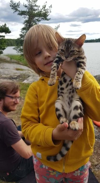 Barn håller upp en liten katt framför en sjö med en vuxen i bakgrunden.