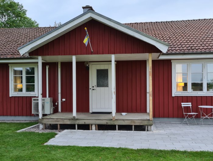 Röd trähusfasad med nyslipade stolpar på verandan, två omålade stolpar, svensk flagga och grönt gräs.