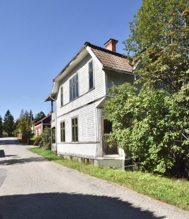 Vitmålat trähus med tegeltak vid en asfalterad väg, med gröna buskar och en klarblå himmel.