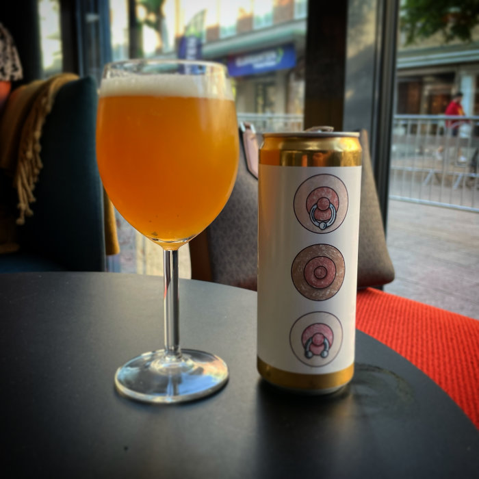 Ett glas med orangefärgad dryck bredvid en ölburk på ett bord, inomhus med utsikt mot gatan.