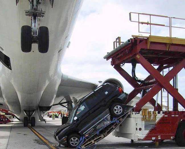 Bil hänger vertikalt från lastutrustning under flygplansnos, misstänkt otillräcklig lastsäkring.