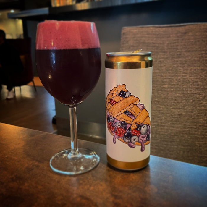 Ett vinglas med mörkröd dryck bredvid en burk med bär och bakverk illustrerade på etiketten.