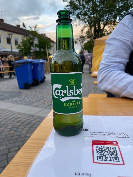En Carlsberg exportflaska på ett bord utomhus med oskarpa människor och tält i bakgrunden.