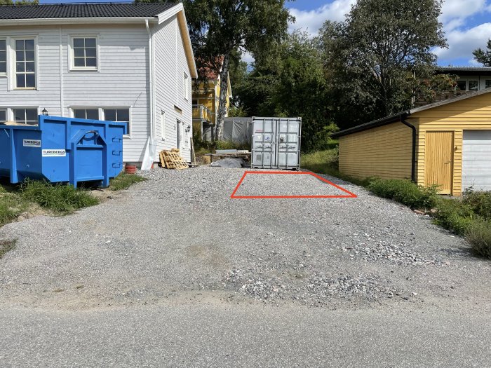 Grusad uppfart med röd markering för planerad carport intill gul garagebyggnad och vit husfasad.