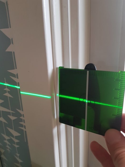 Hand håller ett grönt laserpass mot en vit vägg som projicerar en grön laserlinje, med ojämn tjocklek på linjen.