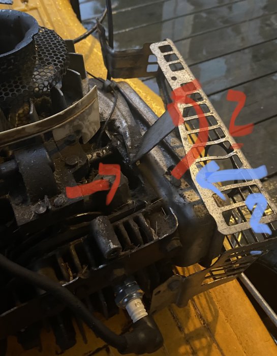 Motorgräsklipparens motor med öppna kåpor och en böjd flärp markerad med röda pilar, nära förgasarens kåpa markerad med blå pil.