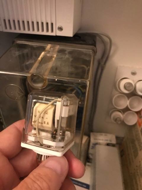 Hand håller ett transparent relä framför en elcentral, med synliga anslutningar och skruvar, relät uppvisar vibrationsskador.