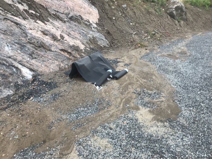 Slänt med grus och lera där leran rann ner i dräneringen, synligt dräneringsrör och geotextil.