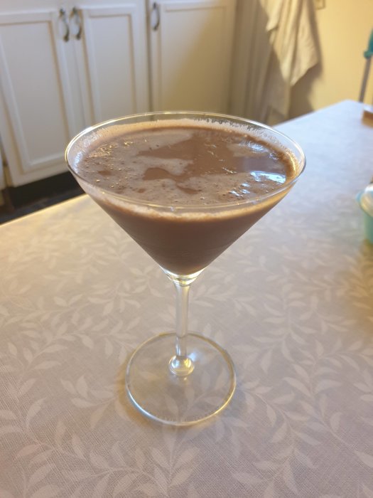 Chokladmartini i cocktailglas på textilklädd bord med köksskåp i bakgrunden.