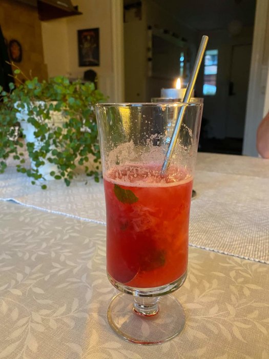 Halvtomt glas med en röd strawberry mojito på ett vitt bord, färgjusterad med karamellfärg, metall sugrör.