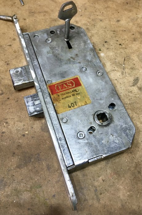 Demonterat FAS407 låskista med trasig fjäder och nyckel, på ett verkstadsbord.