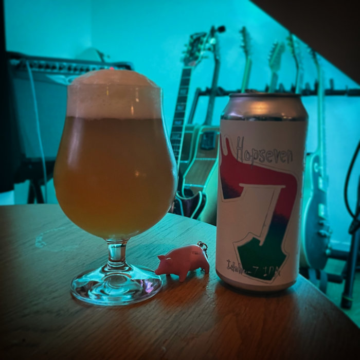 Ölglas med skummande öl bredvid en burk märkt 'Hopseven' och en liten rosa grisfigur, gitarrer i bakgrunden.