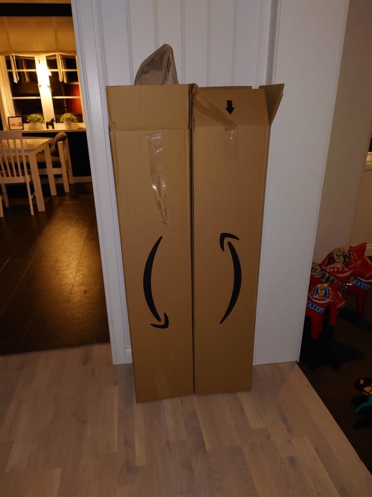 Stort Amazon-paket står på ett trägolv i ett hem mot en vit dörr.