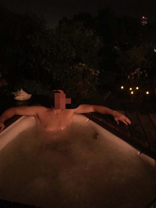 Person som kopplar av i en badtunna utomhus på kvällen med belysning i bakgrunden.