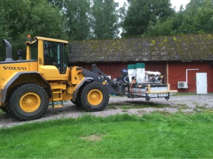 Gul Volvo-hjullastare levererar en CNC-maskin på släpvagn framför röd lada.