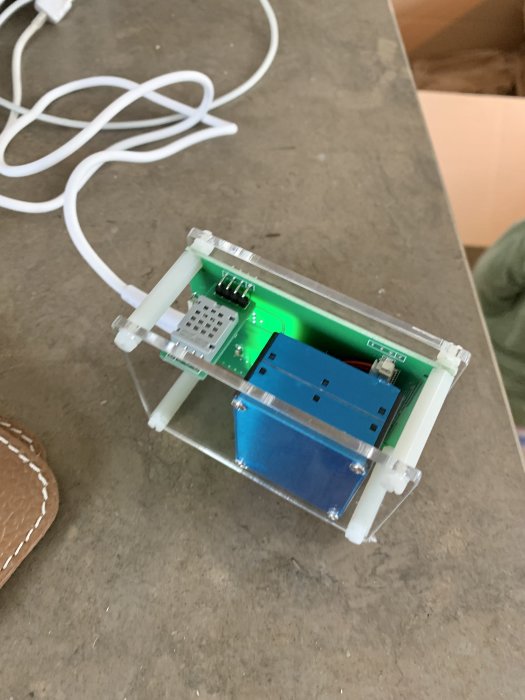 En öppen partikelräknare med blått kretskort och grönt ljus, med en vit USB-kabel, på ett grått bord.