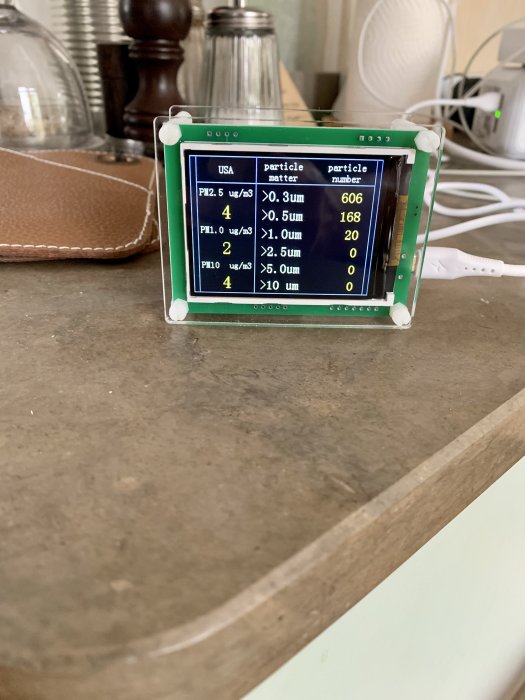 En partikelräknare med digital display som visar PM2.5 och andra partikelstorlekar på en träyta.