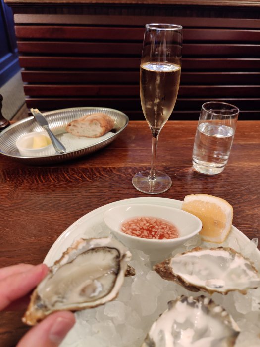 Hand håller ett ostron över ett fat med is och fler ostron, bredvid citron och såsskål. Glas champagne i bakgrunden.