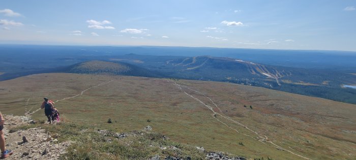 Vy över kargt fjällandskap med vandrare, flacka toppar och skogklädda dalar under klarblå himmel.